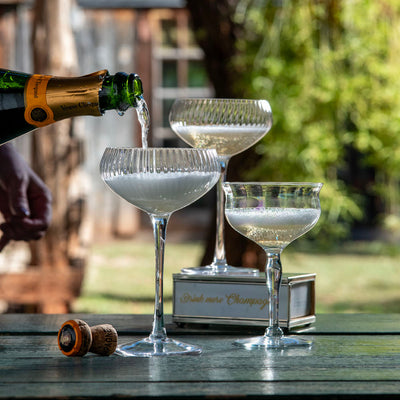A champagne bottle is pouring sparkling wine into three coupe glasses on a wooden table, with a rustic outdoor background of trees and a Perini Ranch Steakhouse in Buffalo Gap, TX.